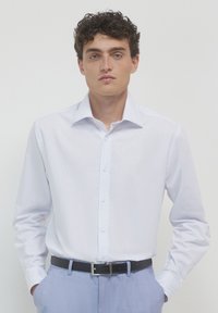 Jeune homme aux cheveux bouclés portant une chemise blanche à rayures fines rentrée dans un pantalon bleu clair avec une ceinture noire, les mains dans les poches.