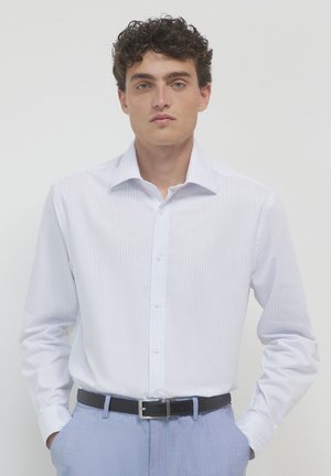 Giovane con capelli ricci che indossa una camicia bianca a righe sottili infilata in pantaloni azzurri chiari con una cintura nera, mani in tasca.