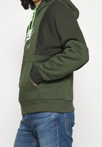 Felpa con cappuccio verde scuro con coulisse e tasca a marsupio. Presenta toppe a contrasto e un logo. Tessuto morbido e strutturato.