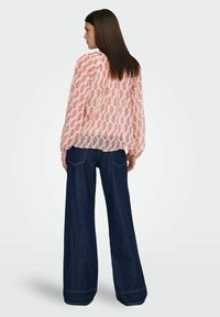 Blouse rose transparente à manches longues avec motif paisley, associée à un jean en denim foncé à jambes larges, présentant une coupe classique et un léger revers à l'ourlet.