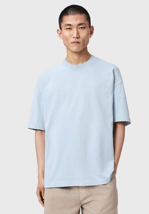 Jeune homme aux cheveux courts portant un T-shirt bleu clair oversize et un pantalon beige, debout devant un fond gris uni.