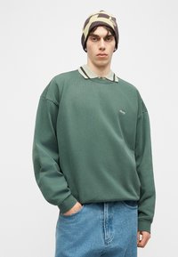 Νέος άνδρας που φοράει πράσινο φαρδύ φούτερ με το λογότυπο «obey», μπλε τζιν, καπέλο με καμουφλάζ, στέκεται μπροστά σε λευκό φόντο.