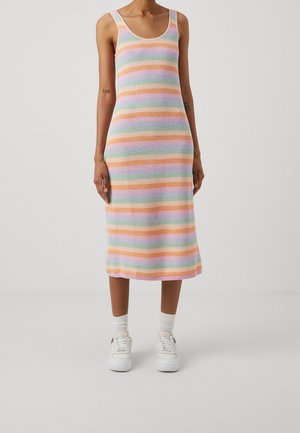 Person trägt ein ärmelloses, knielanges Strickkleid mit horizontalen Pastellstreifen und weiße Sneakers mit Socken, steht vor einem schlichten Hintergrund.