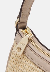 Esquina de un bolso de paja tejido y cuero beige texturizado, con un zipper dorado y una etiqueta, además de una correa de hombro marrón oscuro.