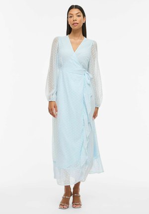 Kvinde iført lyseblå omslagskjole med gennemsigtige lange ærmer og flæsedetalje, stående mod en hvid baggrund.