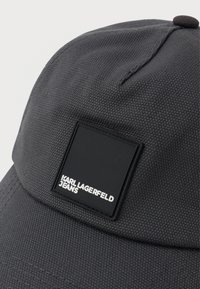 Fekete sapka texturált anyagból, egy téglalap alakú logóval, amelyen a "KARL LAGERFELD JEANS" felirat olvasható fehér színben.