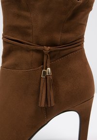 Botte à talon haut en daim marron avec une lanière décorative nouée à pompons autour de la cheville et des embouts métalliques dorés sur les pompons.