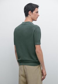 Giovane uomo che guarda a destra, indossando una maglietta a maniche corte di colore verde scuro e pantaloni beige, su uno sfondo chiaro e semplice.