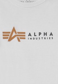 Bijela pamučna majica s velikim smeškim stilizovanim logotipom "A" u smešnoj boji, s tri horizontalne pruge iznad i natpisom "ALPHA INDUSTRIES" ispod.