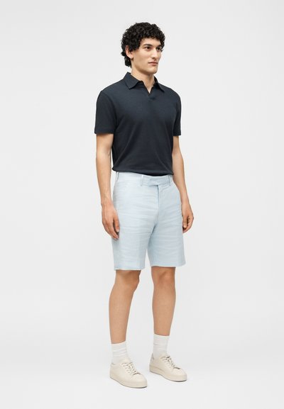 Άνδρας στέκεται φορώντας μαύρο πόλο με κοντά μανίκια, ανοιχτό μπλε σορτς με ζώνη, λευκές κάλτσες και μπεζ αθλητικά παπούτσια.