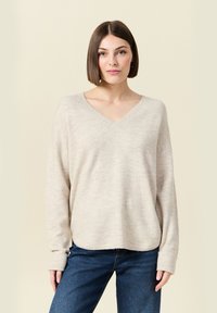 Vrouw met een rechte bruine bob-kapsel, gekleed in een beige V-hals trui en blauwe jeans, staand tegen een lichtbeige achtergrond.