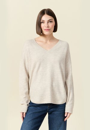 Femme avec un carré lisse brun, portant un pull beige à col en V et un jean bleu, se tenant devant un fond beige clair.