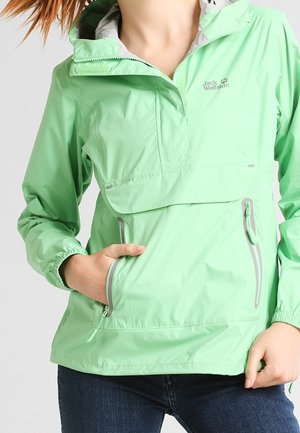 Frau trägt eine hellgrüne Jack Wolfskin Kapuzenjacke mit Reißverschlusstaschen, eine Hand in einer Tasche, vor einfarbigem Hintergrund.