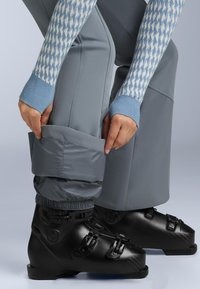 Skidöja i svart med tre justerbara spännen, tillsammans med grå skidbyxor med elastiska mudd och en slät textur.