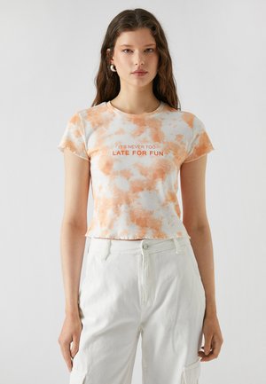 Korte mouwen, cropped t-shirt met een tie-dye patroon in oranje en wit. Tekst leest "HET IS NOOIT TE LAAT VOOR PLEZIER." Zachte, casual stof.