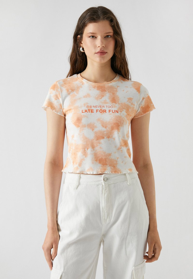 Korte mouwen, cropped t-shirt met een tie-dye patroon in oranje en wit. Tekst leest "HET IS NOOIT TE LAAT VOOR PLEZIER." Zachte, casual stof.