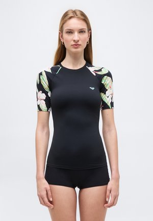 Γυναίκα που φοράει μαύρο μαγιό με κοντά μανίκια, με λουλουδάτο σχέδιο στα μανίκια και μαύρο μαγιό σορτς που ταιριάζει, στέκεται μπροστά σε απλό φόντο.