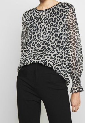 Woman wearing a long-sleeve sheer blouse with black and white leopard print and black fitted pants, standing against a plain background.
