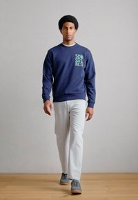 Tmavomodrá mikina so zelenou grafikou na hrudi, kombinovaná s bielymi nohavicami a sivými teniskami, pózujúca na jednoduchom pozadí.