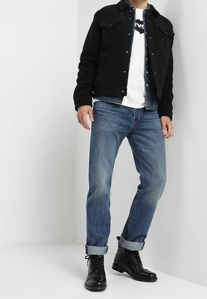 Homme portant une veste sherpa noire, un t-shirt graphique blanc, un jean bleu avec revers et des bottes en cuir noires à lacets, debout contre un mur blanc.