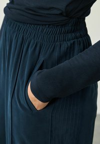 Persoon in donkerblauwe elastische broek met zijzakken, hand in de zak gestoken, gecombineerd met een donkerblauw shirt met lange mouwen.