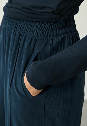 Persoană îmbrăcată cu pantaloni elastic aținti de culoare albastră închisă, cu buzunare laterale, cu mâna introdusă în buzunar, asortați cu un tricou cu mâneci lungi de culoare albastră închisă.