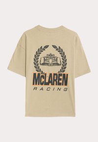 Beige tričko s grafickým potiskem, na kterém je závodní auto a vavřínový věnec. Černý a oranžový text "McLaren Racing" ve spodní části. Krátké rukávy.