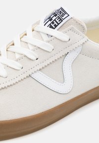 Gros plan sur une basket en suède beige avec des lacets blancs, une bande latérale en cuir blanc, une semelle en gomme et le logo Vans sur l’étiquette de la languette.