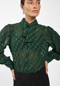 Groene, doorzichtige blouse met een gestructureerd zigzagpatroon, striknek en knoopsluiting aan de voorkant. Lange mouwen met geplooide manchet.
