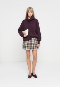Pull violet surdimensionné avec un col montant, associé à une mini-jupe à carreaux dans des tons bruns et crémeux, et des ballerines bordeaux brillantes.