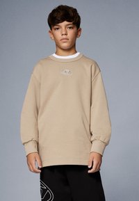 Ragazzo con i capelli castani corti che indossa una camicia beige a maniche lunghe e pantaloni neri con un logo bianco su uno sfondo grigio uniforme.