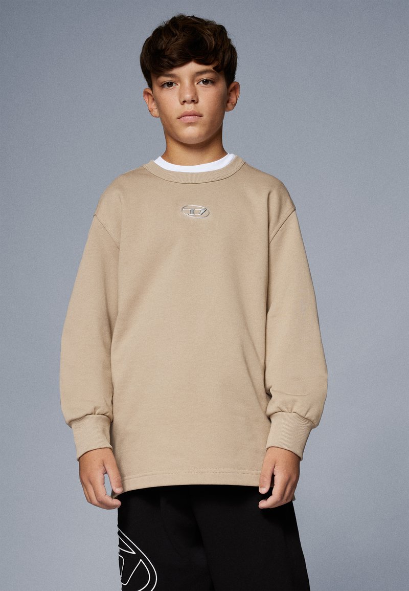 Ragazzo con i capelli castani corti che indossa una camicia beige a maniche lunghe e pantaloni neri con un logo bianco su uno sfondo grigio uniforme.