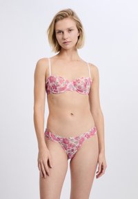 Ensemble bralette et string en dentelle rose avec un motif floral, présentant un tissu transparent, des épaules délicates et un petit nœud sur le string.