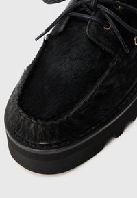 Zwarte schoen met een vachttextuur en een ronde neus, gedetailleerde stiksels en zwarte veters. De schoen heeft een stevige zwarte rubberen zool met een gemarmerd profiel.