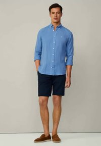 Lysblå linned skjorte med lange ærmer og knaplukning, parret med marineblå shorts og brune slip-on sko. Glat tekstur og afslappet pasform.