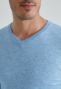 Pull à col en V bleu clair, fabriqué à partir d'un matériau doux, avec une texture subtile et des bords côtelés. Gros plan sur l'encolure et le haut du corps.