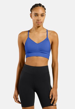Femme portant un brassière de sport bleue et un short cycliste taille haute noir, debout avec une expression neutre sur un fond blanc.