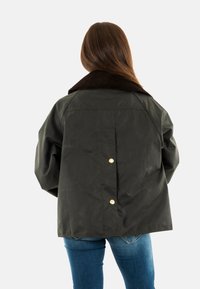 Dunkelgrüne Jacke mit einem Cordkragen, oversized Schnitt und goldenen Druckknöpfen an der Rückseite. Glatte Textur mit leichtem Glanz.