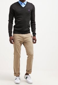 Pull noir à col en V sur une chemise à col bleu clair, assorti à un chino kaki et des baskets blanches avec des accents verts.