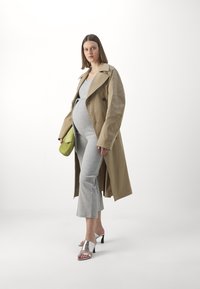 En tan trenchcoat over en grå jumpsuit med vidde bukser, kombinert med sølv sandaler og en grønn veske. Glatte teksturer og lagdelt design.