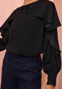 Blusa nera con maniche a volant e dettaglio a fiocco sul collo, abbinata a jeans scuri in denim con due tasche posteriori.