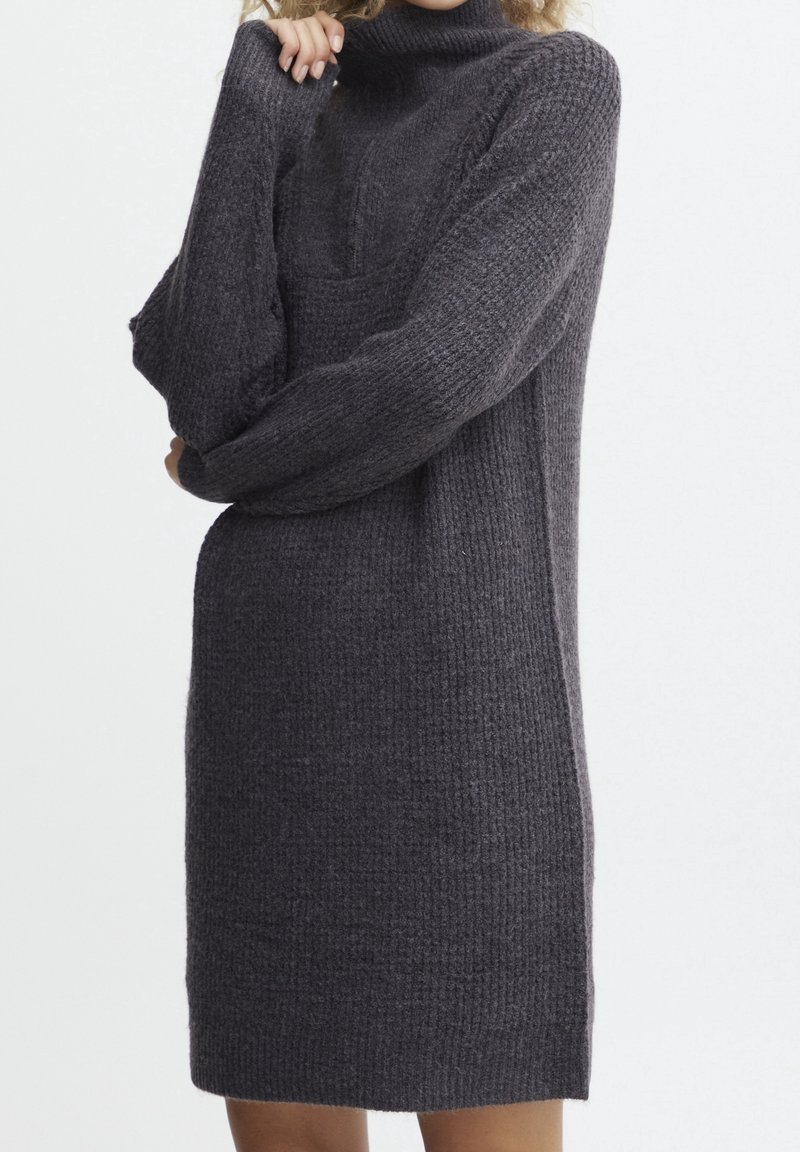 Femme portant une longue robe pull en maille gris foncé à col roulé et manches longues, debout devant un fond blanc.