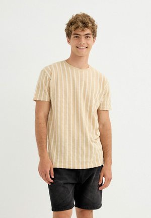 Joven con cabello rizado, vestido con una camiseta beige de rayas verticales y pantalones cortos de mezclilla negros, sonriendo frente a un fondo blanco liso.