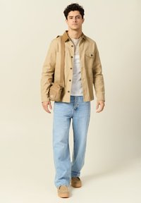 Vaaleanruskea nappikauluksellinen takki valkoisen kuviollisen paidan päällä, yhdistettynä vaaleansinisiin farkkuihin ja vaaleanruskeisiin kenkiin. Mukana pieni vaaleanruskea olkalaukku.