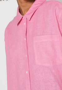 Pink linen button-up shirt with a classic collar, featuring a single chest pocket and a textured fabric. Buttons are white.