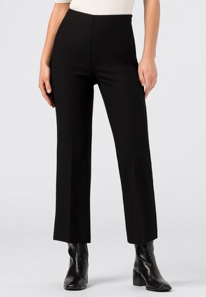 Vrouw draagt rechte zwarte hoge taillebroek gecombineerd met zwarte enkellaarsjes met hak op een lichte achtergrond.