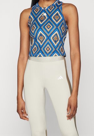 Žena ve modrém a hnědém geometrickém krátkém topu bez rukávů a světle béžových legínách s vysokým pasem, s bílým logem Adidas na levém stehně.