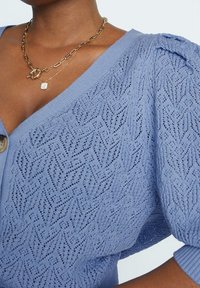 Jasnoniebieski sweter z dzianiny z wyraźnym wzorem w diamenty, głębokim dekoltem w kształcie V oraz puffowanymi krótkimi rękawami.
