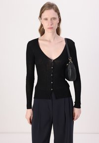 Schwarze gerippte Langarm-Cardigan mit tiefem V-Ausschnitt und Knopfverschluss. Begleitet von einer kleinen schwarzen Tasche, getragen über einer marineblauen Hose.