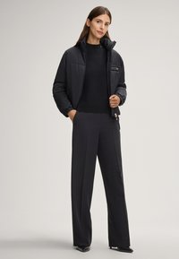 Schwarze Pufferjacke mit hohem Kragen, Reißverschlusstasche, kombiniert mit einem schwarzen Strickpullover und weitem Schnitt bei den Hosen. Glattes Material, taillierte Passform.